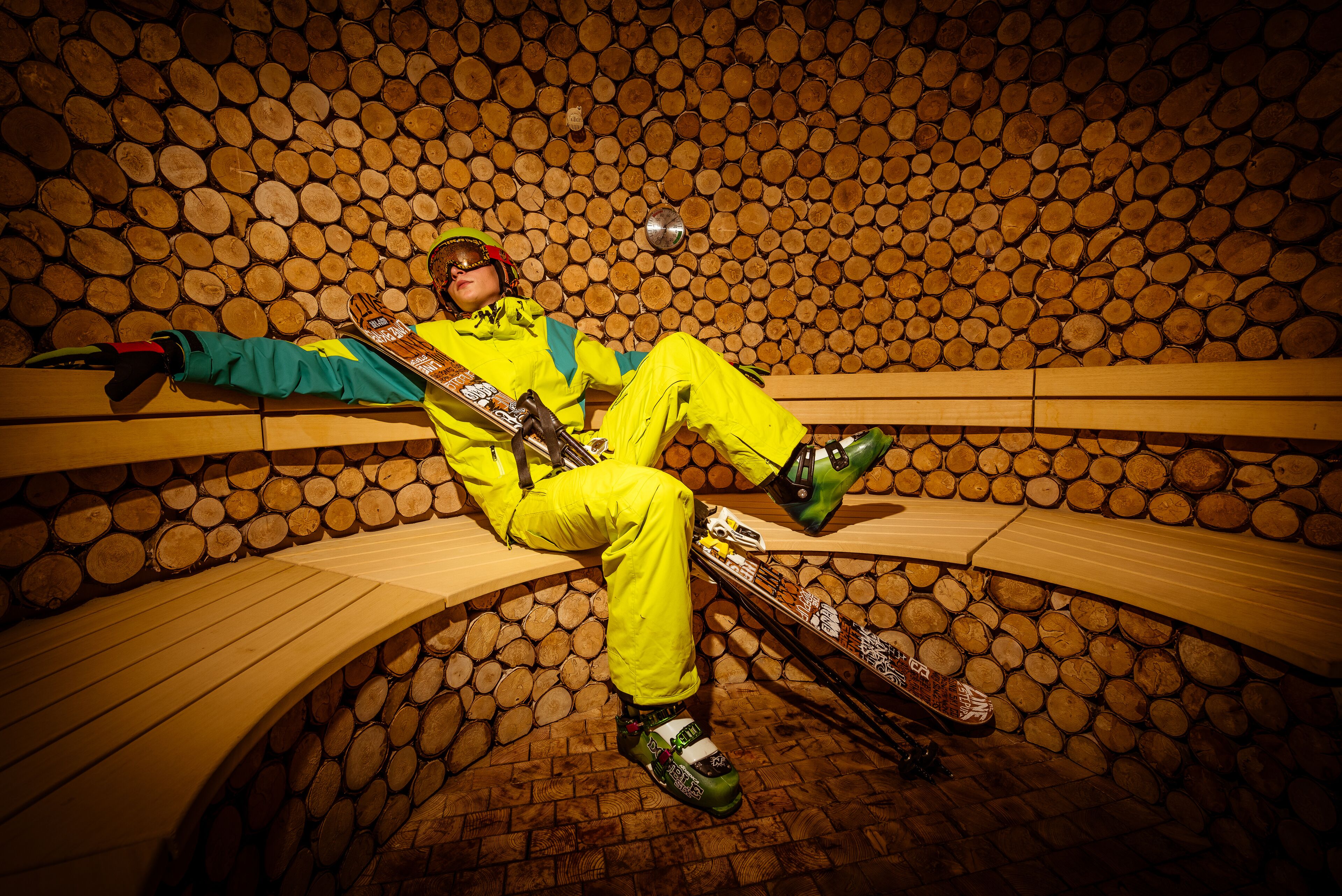Une personne vêtue d’une combinaison de ski jaune vif est assise sur un banc en bois dans une pièce dont les murs sont faits de rondins de bois empilés.