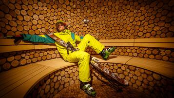 Una persona con equipo de esquí amarillo brillante sentada en un banco de madera en una habitación con paredes construidas con troncos apilados.