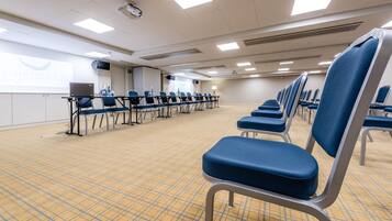 Een congreszaal met rijen blauwe stoelen, een scherm voor een beamer en een laptop op een tafel.