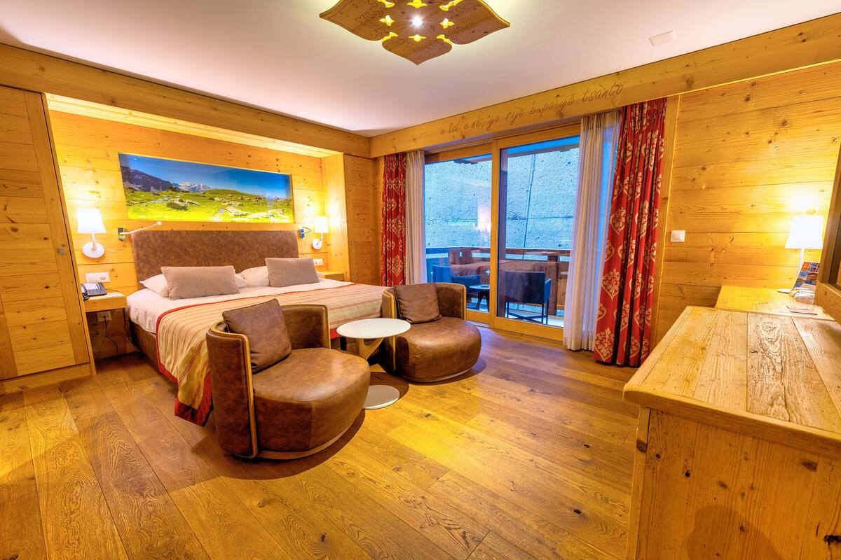 Habitación amueblada con madera, con cama, dos sillones, una mesita y un ventanal con cortinas.