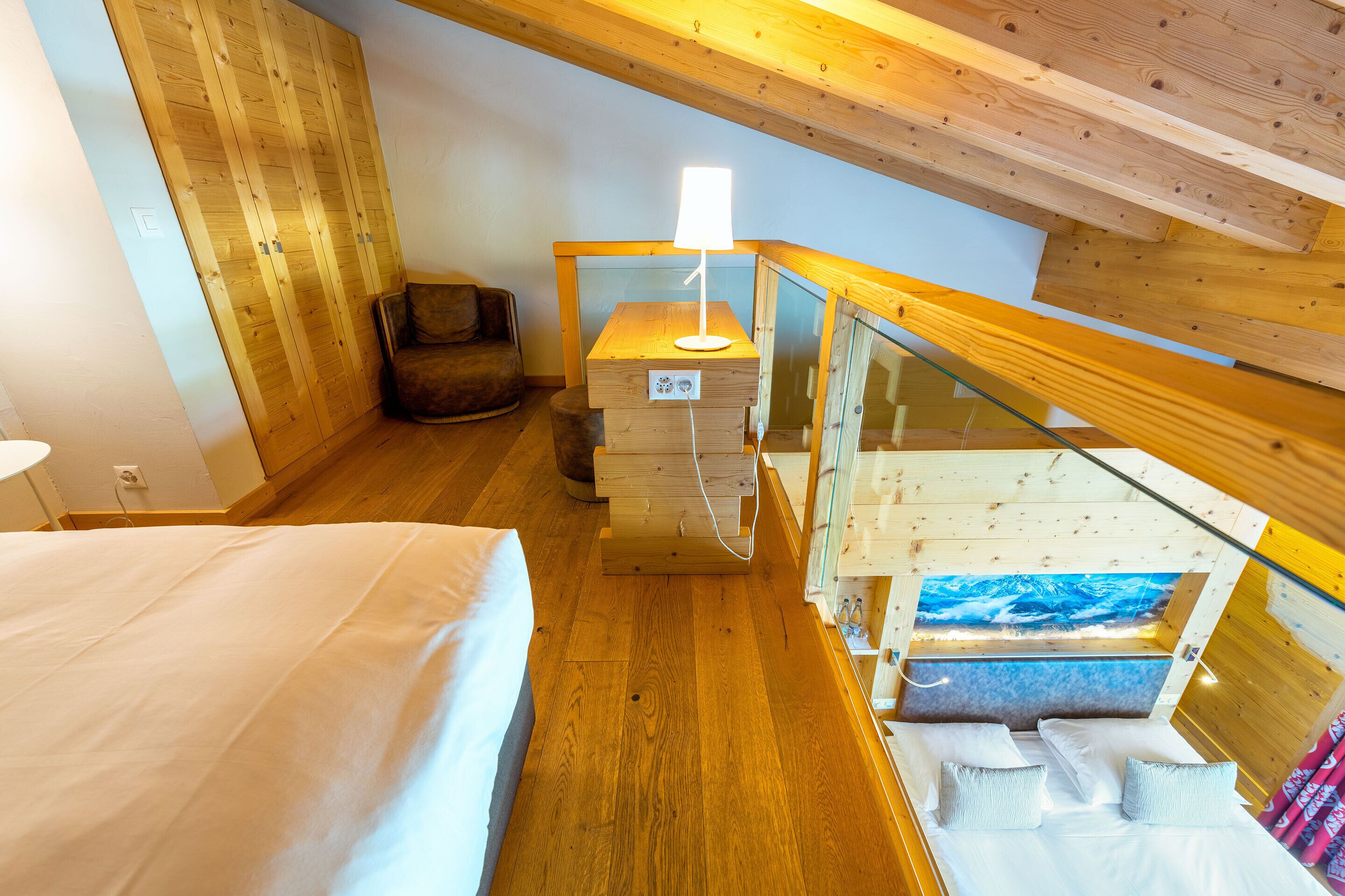 A bedroom with a bed, a nightstand, a lamp, a chair, and a glass railing.