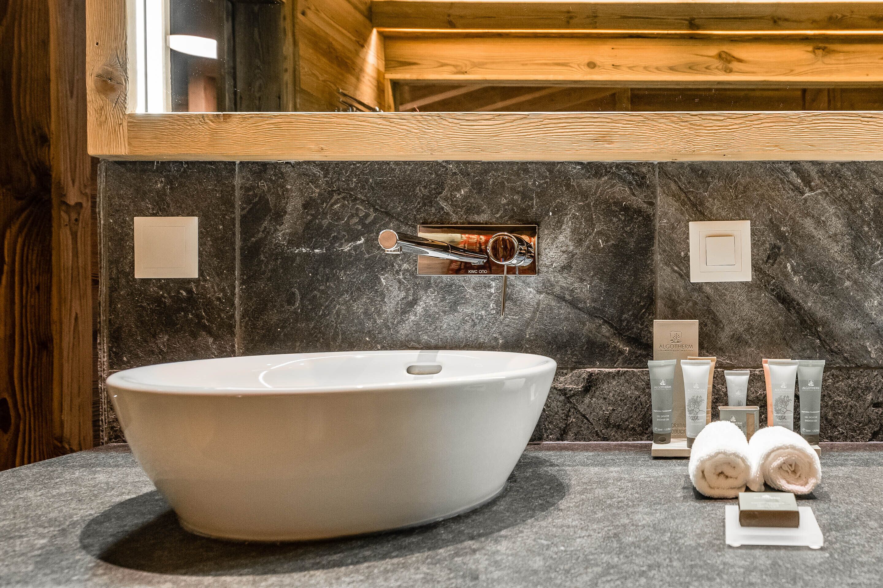 Un baño moderno con un lavamanos ovalado grande, paredes de mármol oscuro, vigas de madera y un espejo arriba.