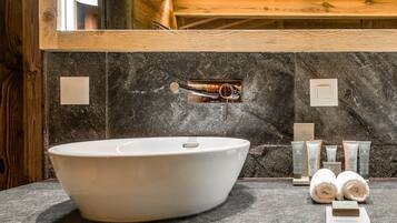 Un baño moderno con un lavamanos ovalado grande, paredes de mármol oscuro, vigas de madera y un espejo arriba.