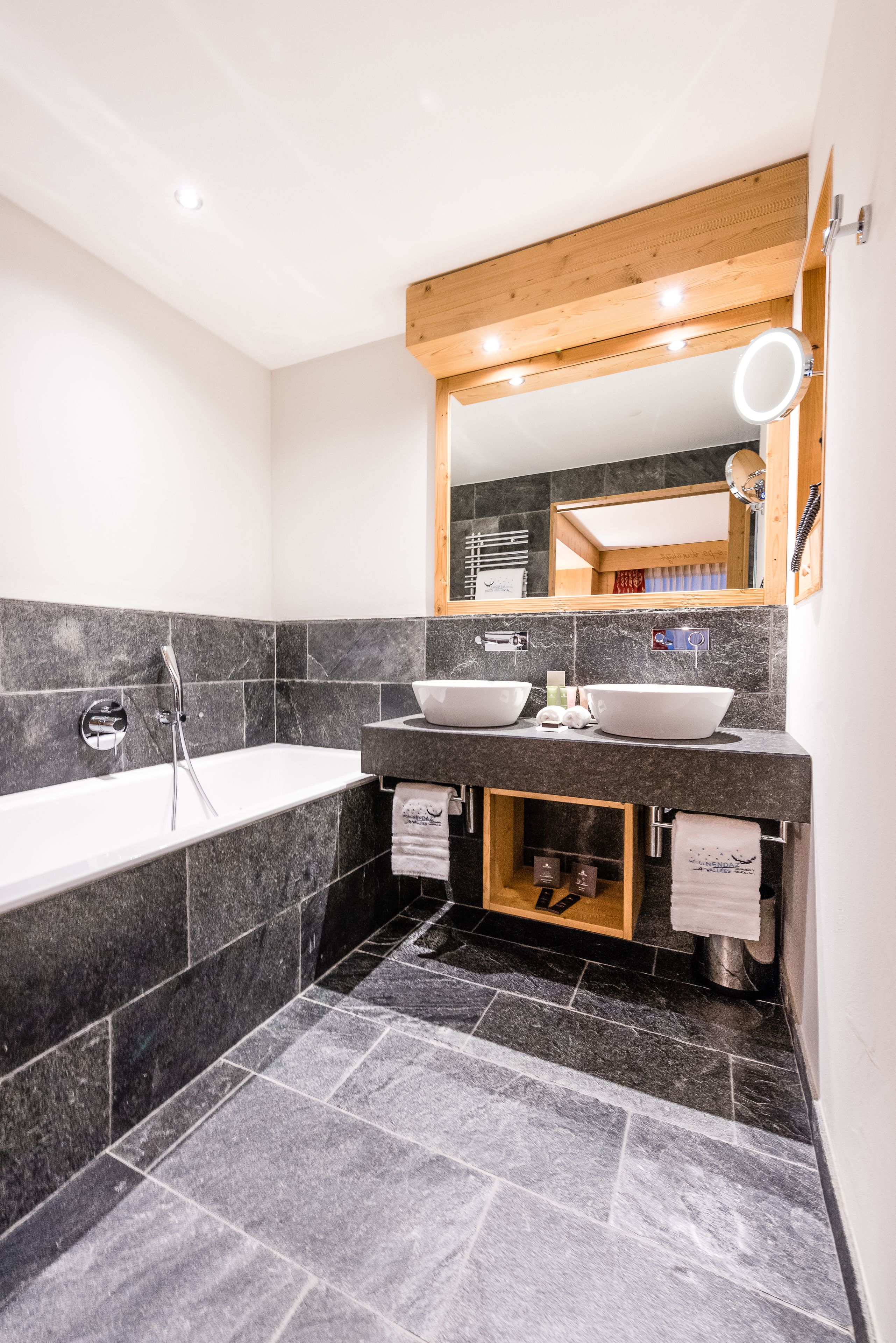 Une salle de bain moderne avec une baignoire, deux lavabos, un meuble-lavabo en bois et un miroir avec éclairage intégré.