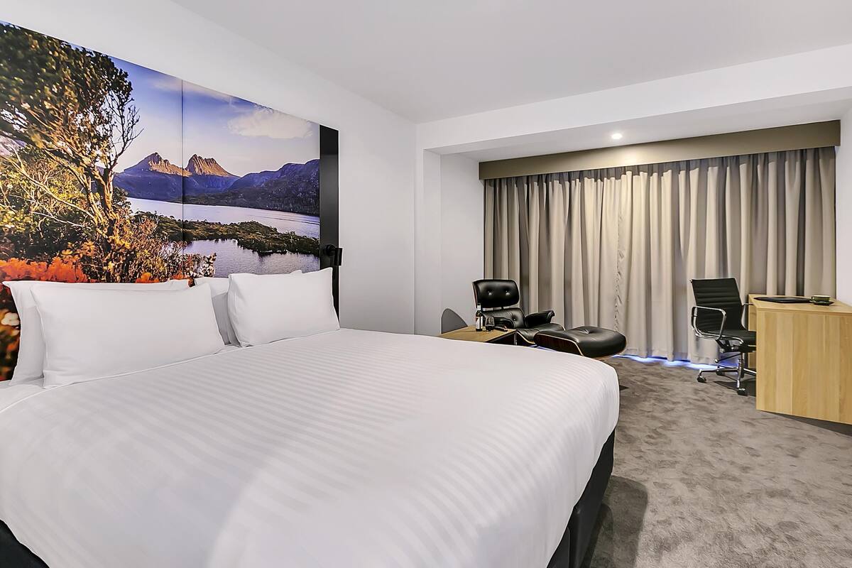 A hotel room with a large bed, a desk, a chair, and a view of mountains and a lake.