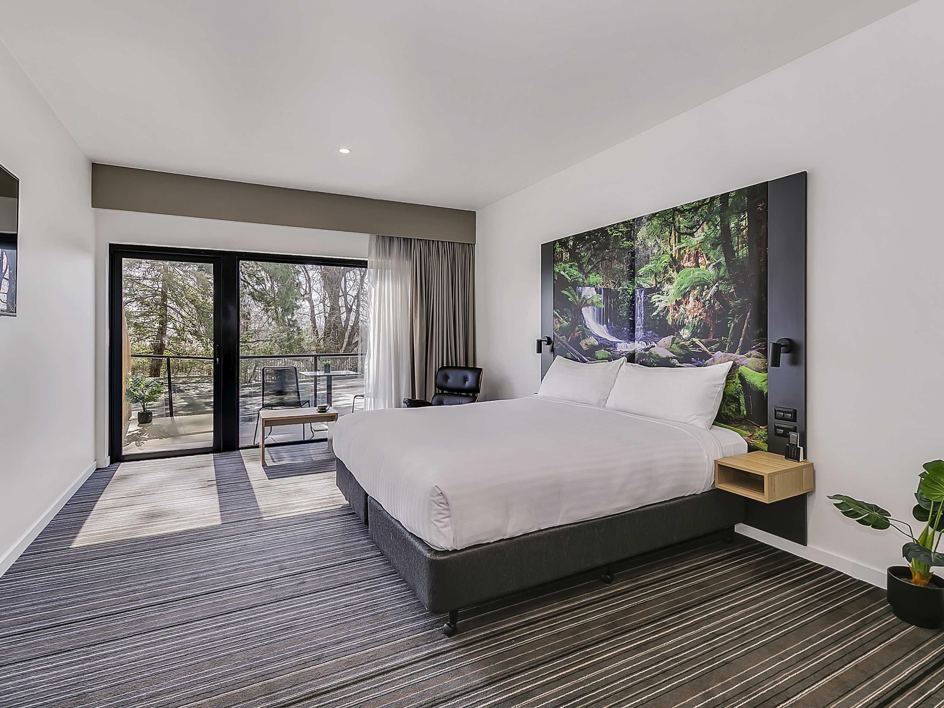 A modern hotel room with a large bed, a view of the outdoors, a dining area, and a large painting on the wall.