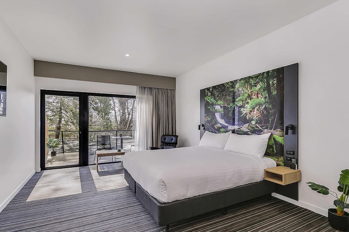 A modern hotel room with a large bed, a view of the outdoors, a dining area, and a large painting on the wall.