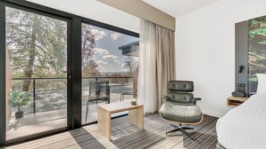 Una habitación de hotel moderna con un amplio ventanal corredero que da a un balcón, un sillón cómodo, una mesita y una cama.