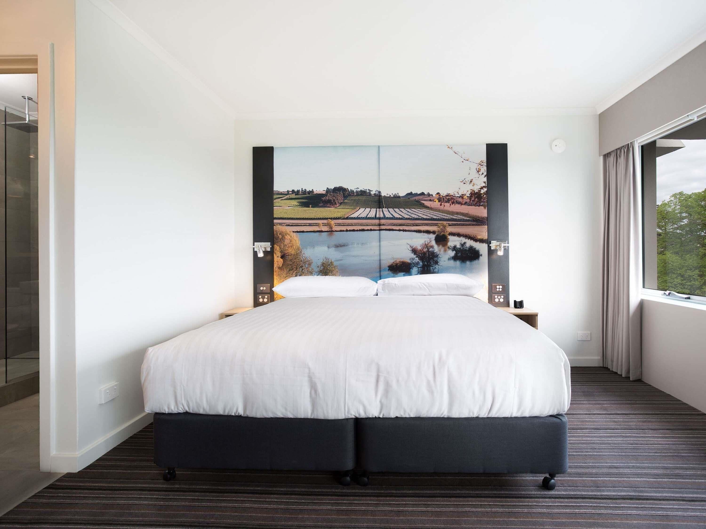 Een moderne hotelkamer met een groot bed, uitzicht op een landschap door een raam en een bedieningspaneel aan de muur.