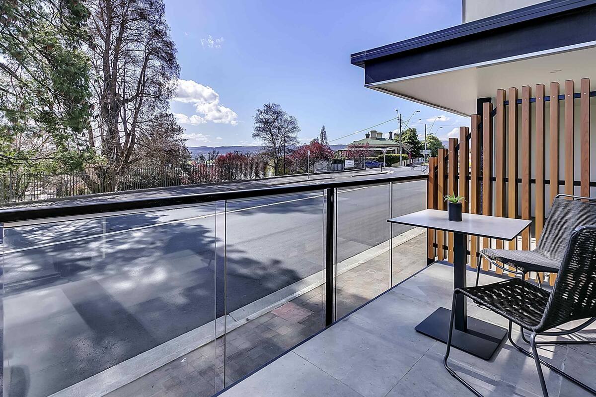 A balcony with a glass railing, a table with a chair, and a view of a road and trees.