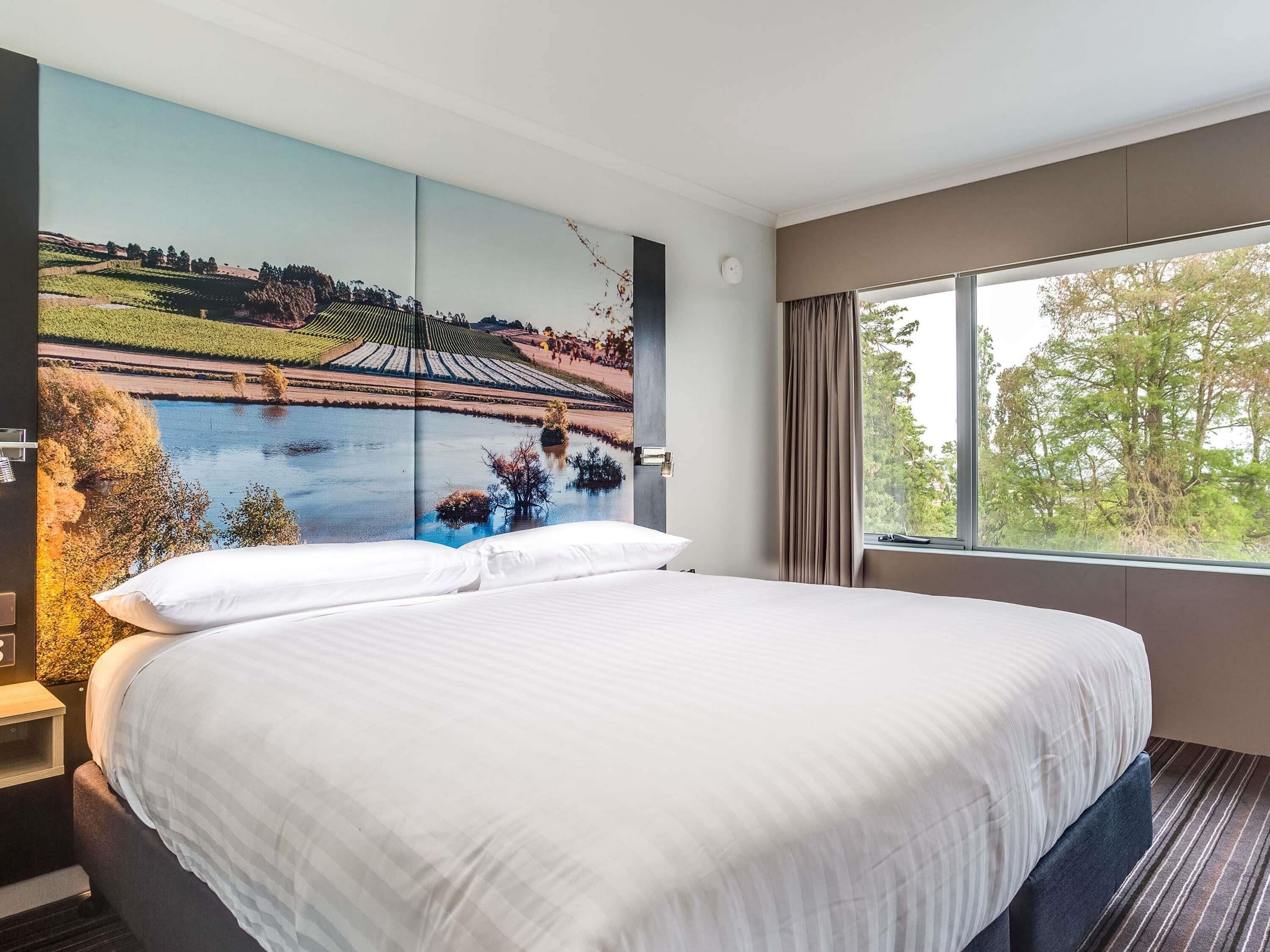 A hotel room with a large bed, a view of a landscape painting, a window with curtains, and a nightstand with a lamp.