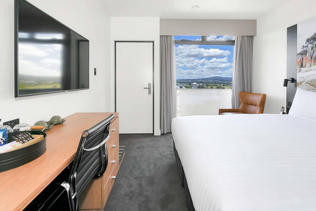 A hotel room with a bed, a desk, a chair, a TV, a door, and a window with a view.