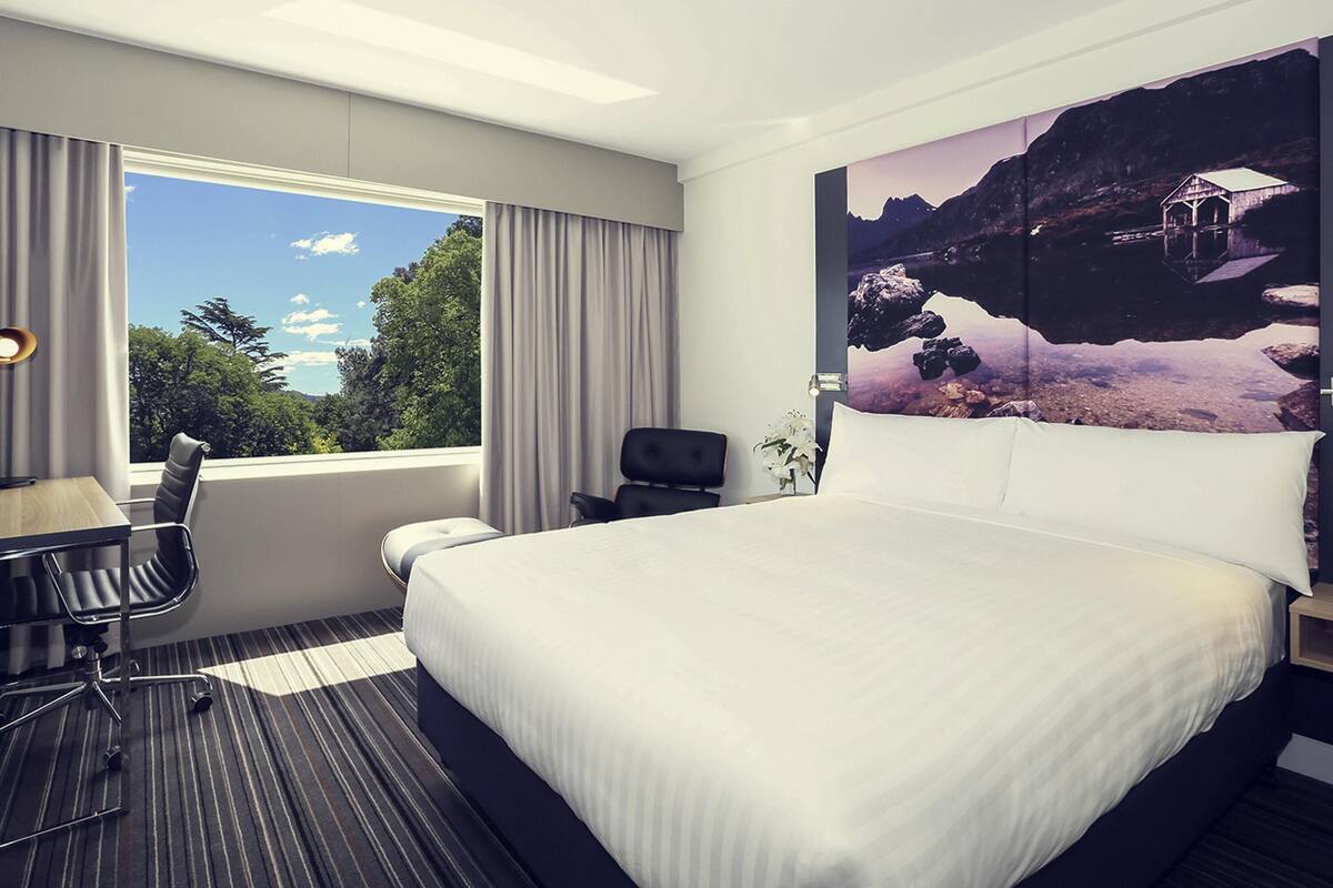 A hotel room with a large bed, a desk, and a chair. There is a window with curtains, and a painting of a landscape on the wall.
