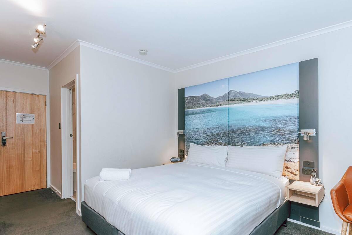 A bedroom with a bed, a nightstand, a chair, a door, and a wall with a scenic painting.