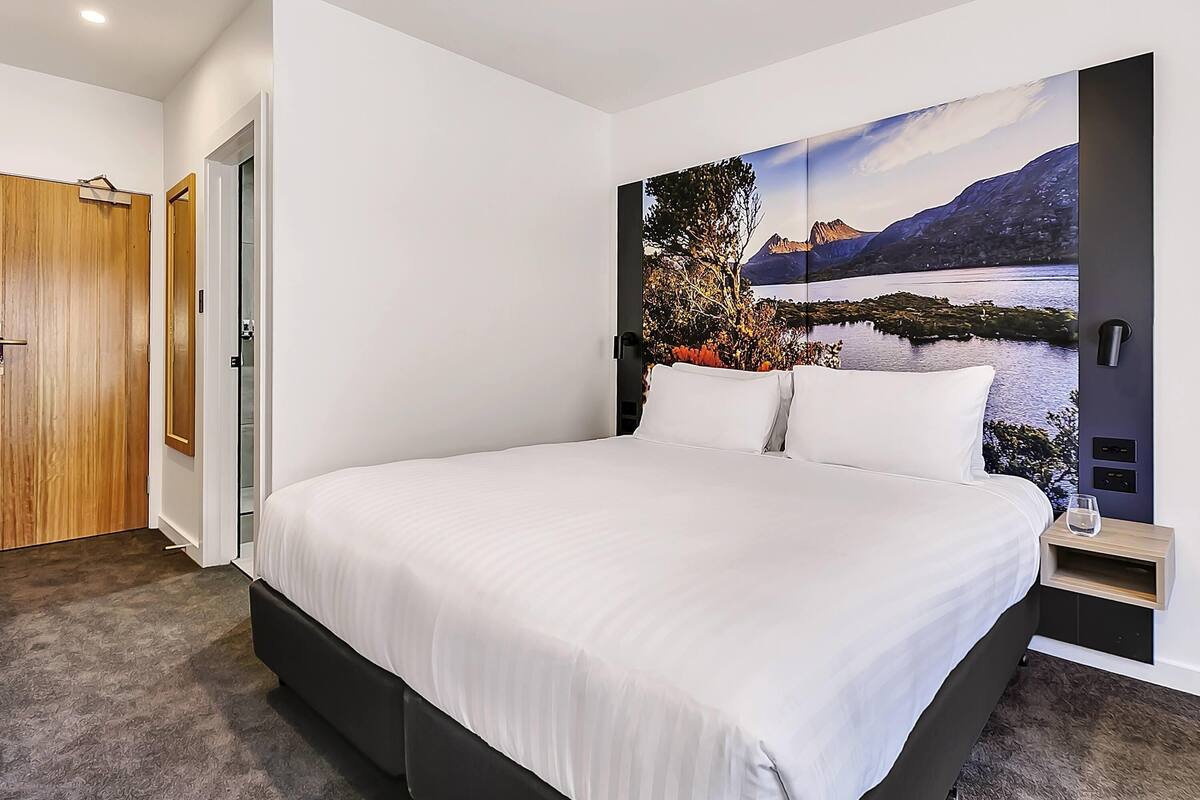 A modern bedroom with a large bed, a wooden door, a mirror, and a scenic wall mural.