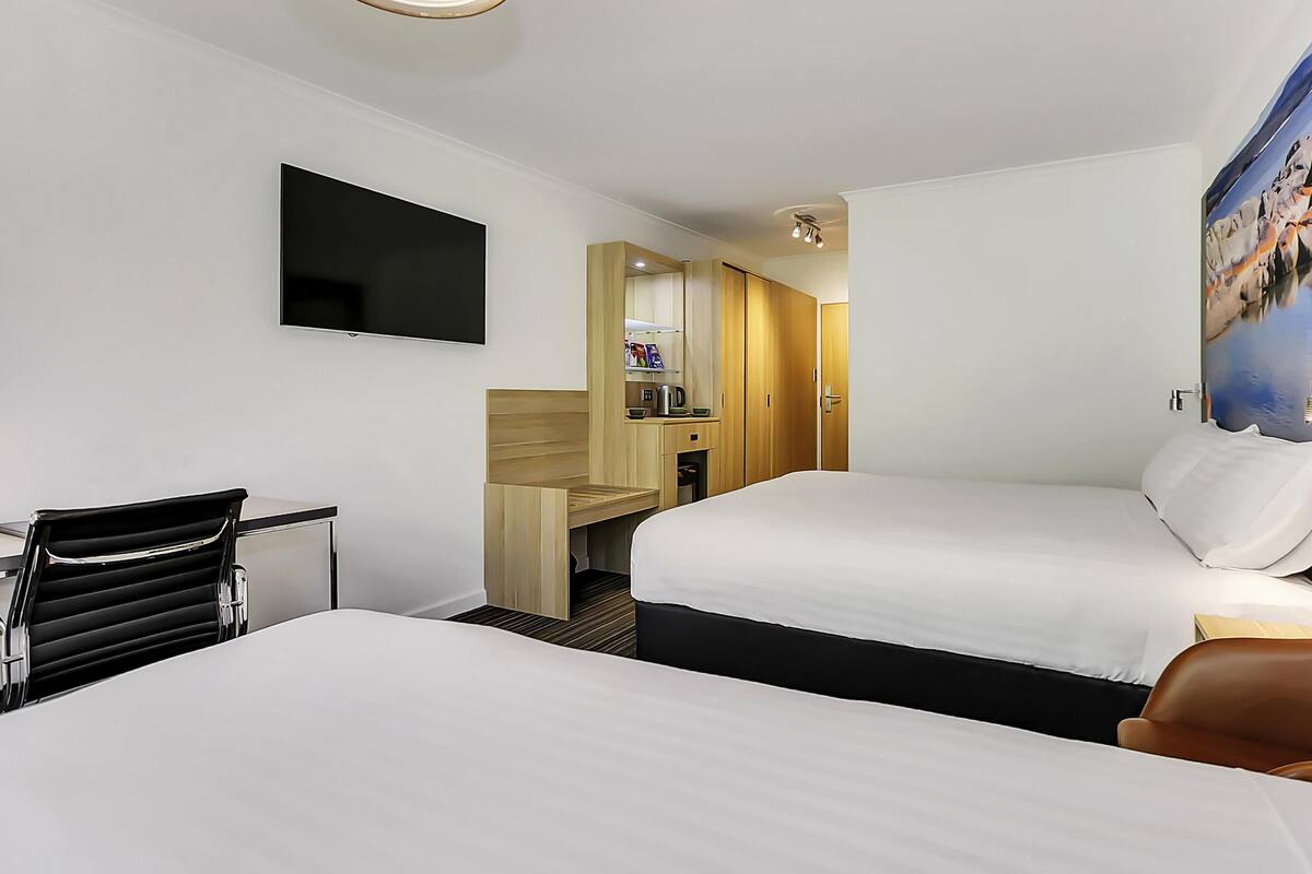 A hotel room with two beds, a desk, a chair, a TV, a mirror, and a painting on the wall.