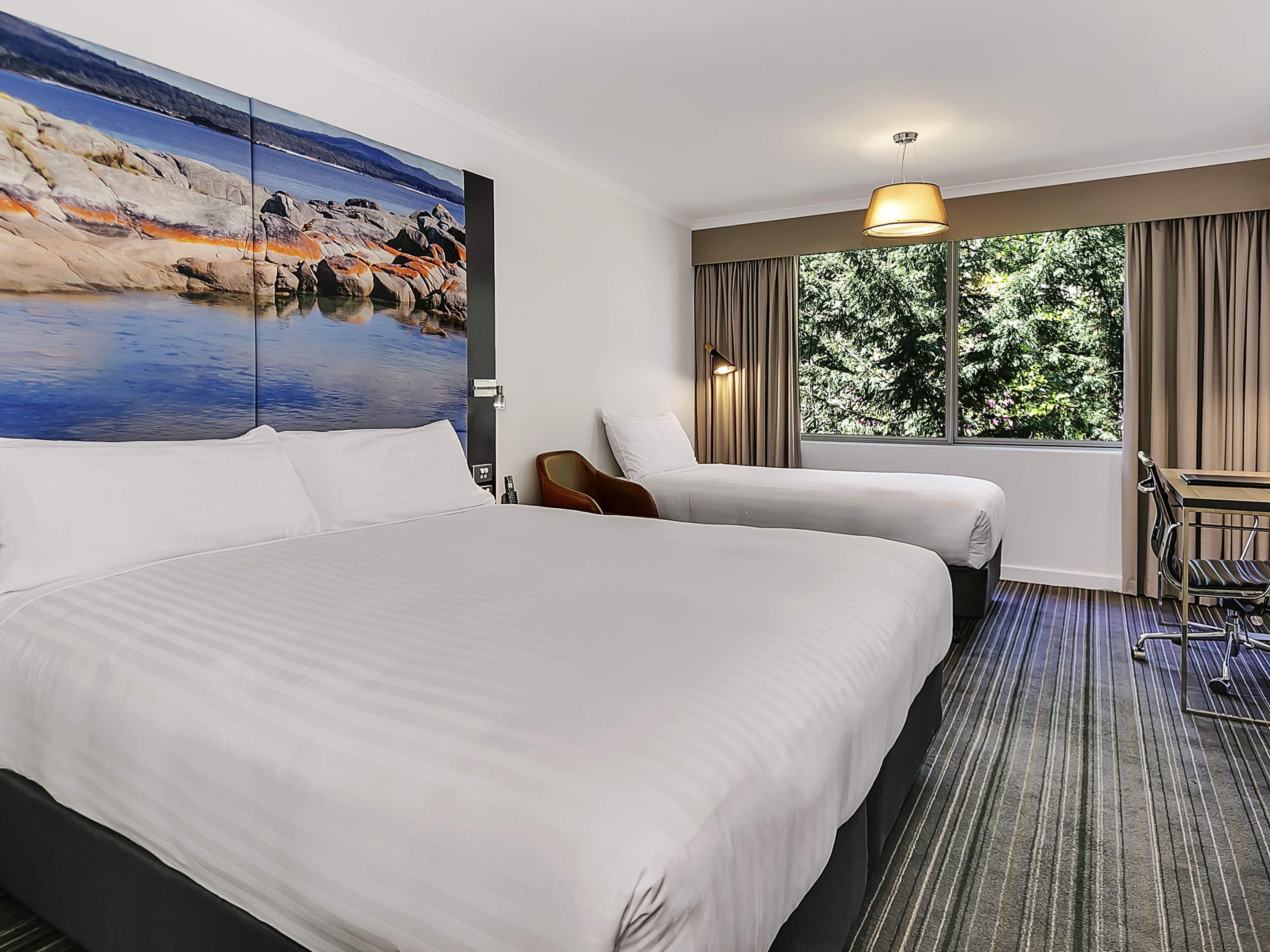 A hotel room with a large bed, a desk, a chair, and a window with a view of trees.
