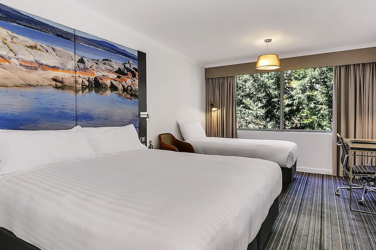 A hotel room with a large bed, a desk, a chair, and a window with a view of trees.