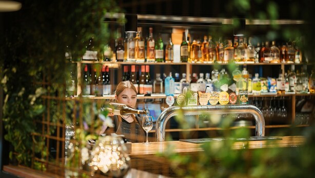 Zona de bar con estantes llenos de botellas y una persona sirviendo una bebida.