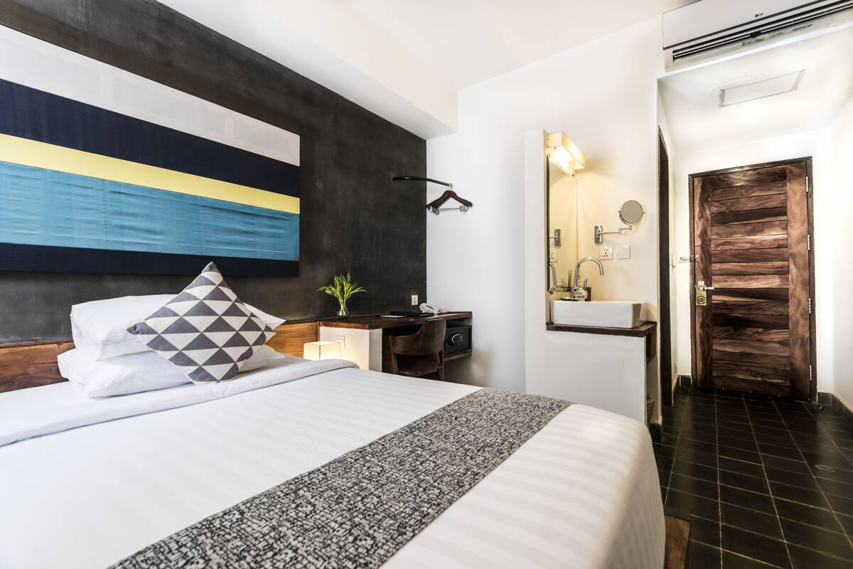 Ein modernes Hotelzimmer mit einem großen Bett, einem Nachttisch mit Lampe, einem Schreibtisch mit Stuhl, einem Spiegel und einer Holztür.
