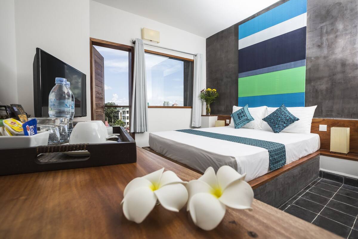 Ein Hotelzimmer mit Bett, Schreibtisch mit Tablett, Wasserflasche und Blumen in einer Vase.