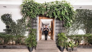 Una persona está de pie frente a la entrada de un edificio con puerta de madera y reja de metal, rodeado de abundante vegetación y plantas en macetas.
