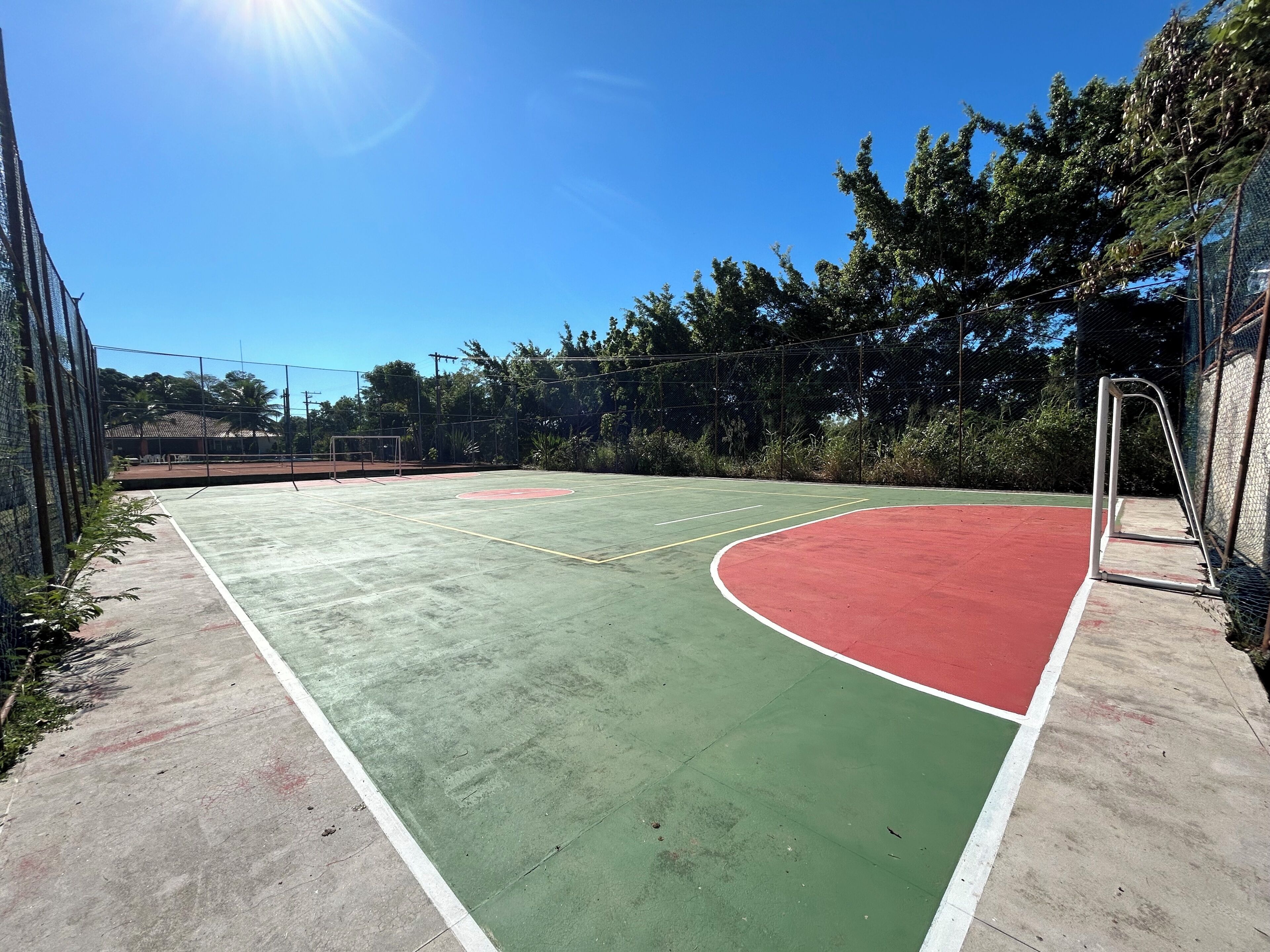 Un terrain de basket en plein air, avec une surface de jeu verte et rouge, entouré d’une clôture en treillis métallique et d’arbres.