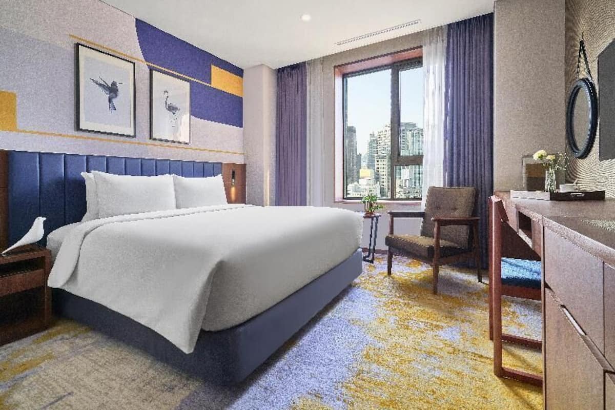 Habitación de hotel con cama, escritorio, silla, ventana con vista a la ciudad y cuadros en la pared.
