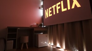 A room with a large screen displaying the word "NETFLIX" in bold letters, a small desk with a computer, a chair, and a window with curtains. - OYO Hostel Myeongdong 2 (Seoul)