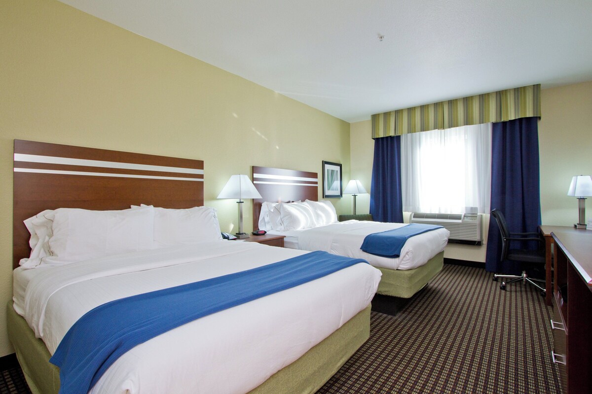 Habitación de hotel con dos camas, un escritorio, una silla y un ventanal con cortinas.
