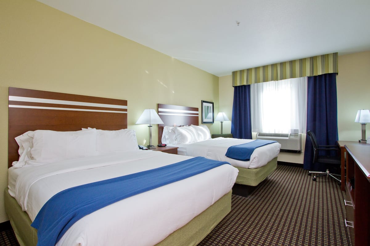 Une chambre d’hôtel avec deux lits, un bureau, une chaise, une lampe et une fenêtre avec des rideaux.