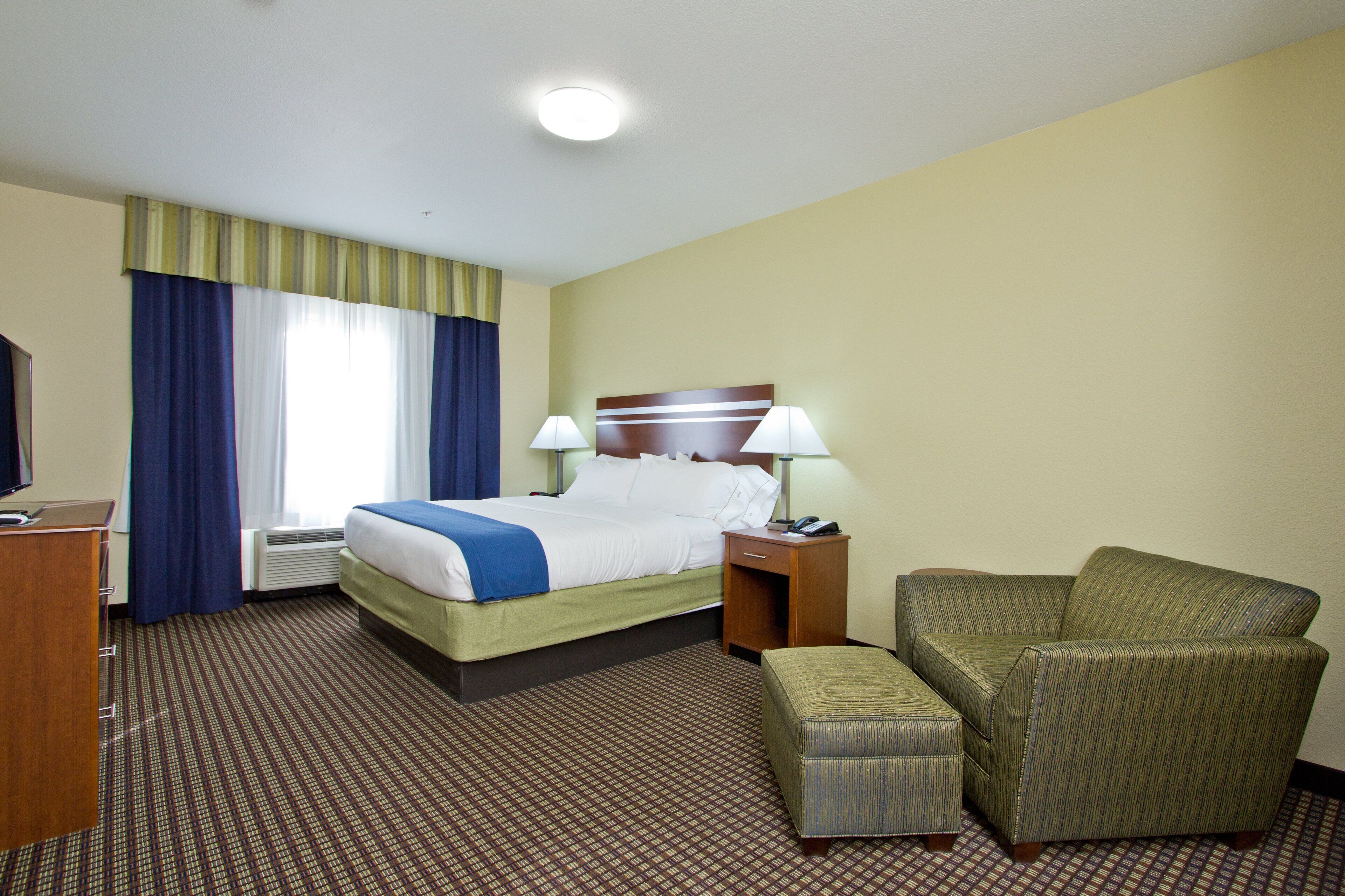 A hotel room with a bed, a chair, an ottoman, a desk, and a television.