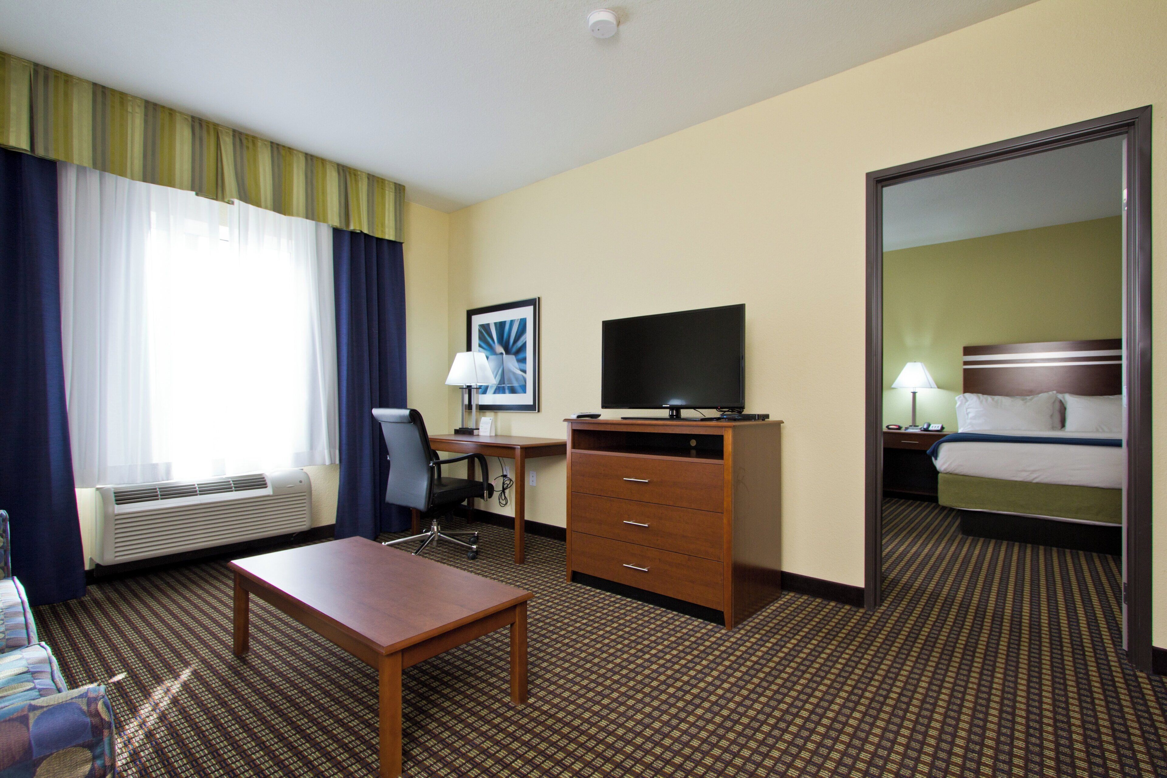 A hotel room with a bed, a desk with a chair, a television, a dresser, and a window with curtains.