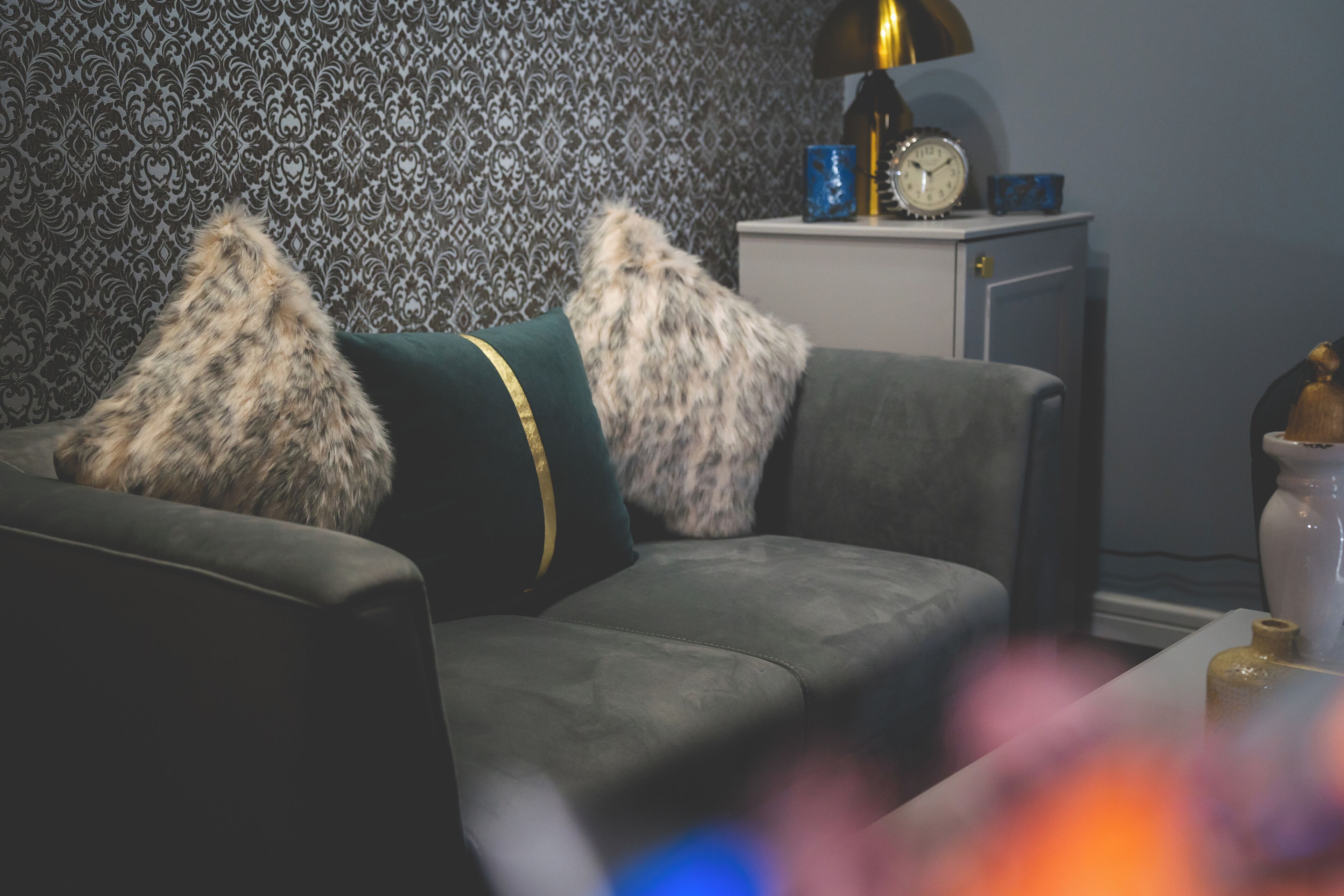A living room with a sofa, patterned wallpaper, a lamp, a clock, and a cabinet.
