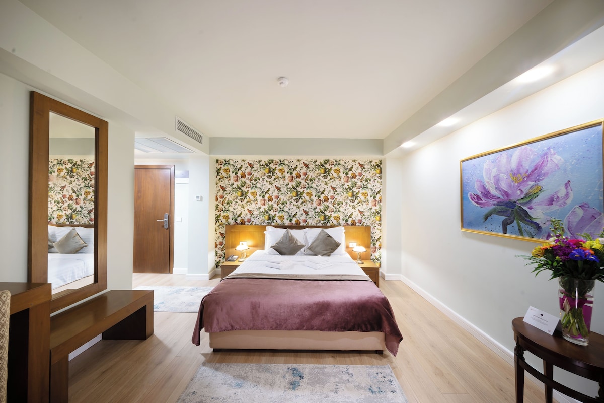 A bedroom with a bed, bedside lamps, a mirror, a wooden bench, a floral wallpaper, a painting, and a vase with flowers.