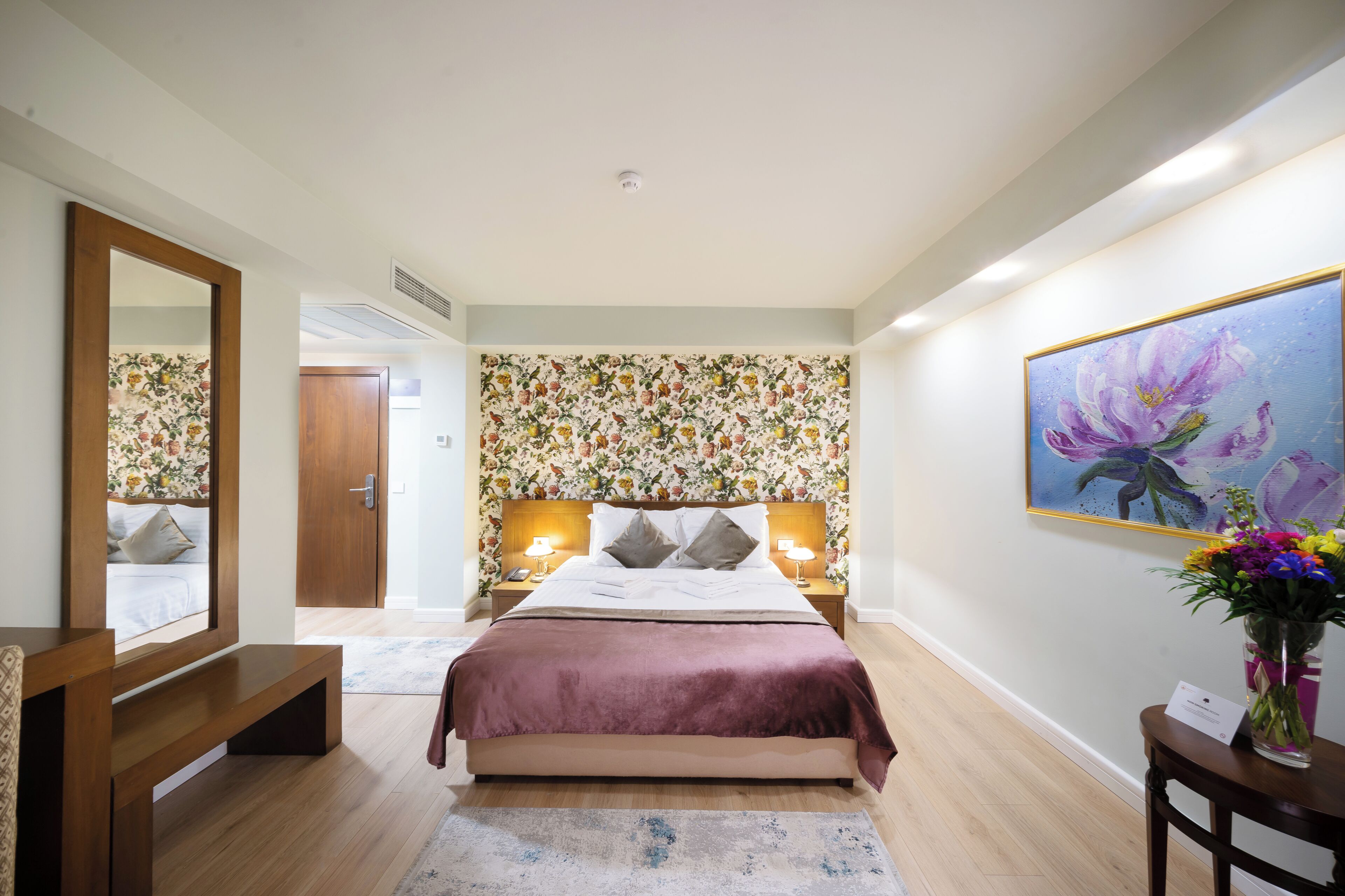 A bedroom with a bed, bedside lamps, a mirror, a wooden bench, a floral wallpaper, a painting, and a vase with flowers.