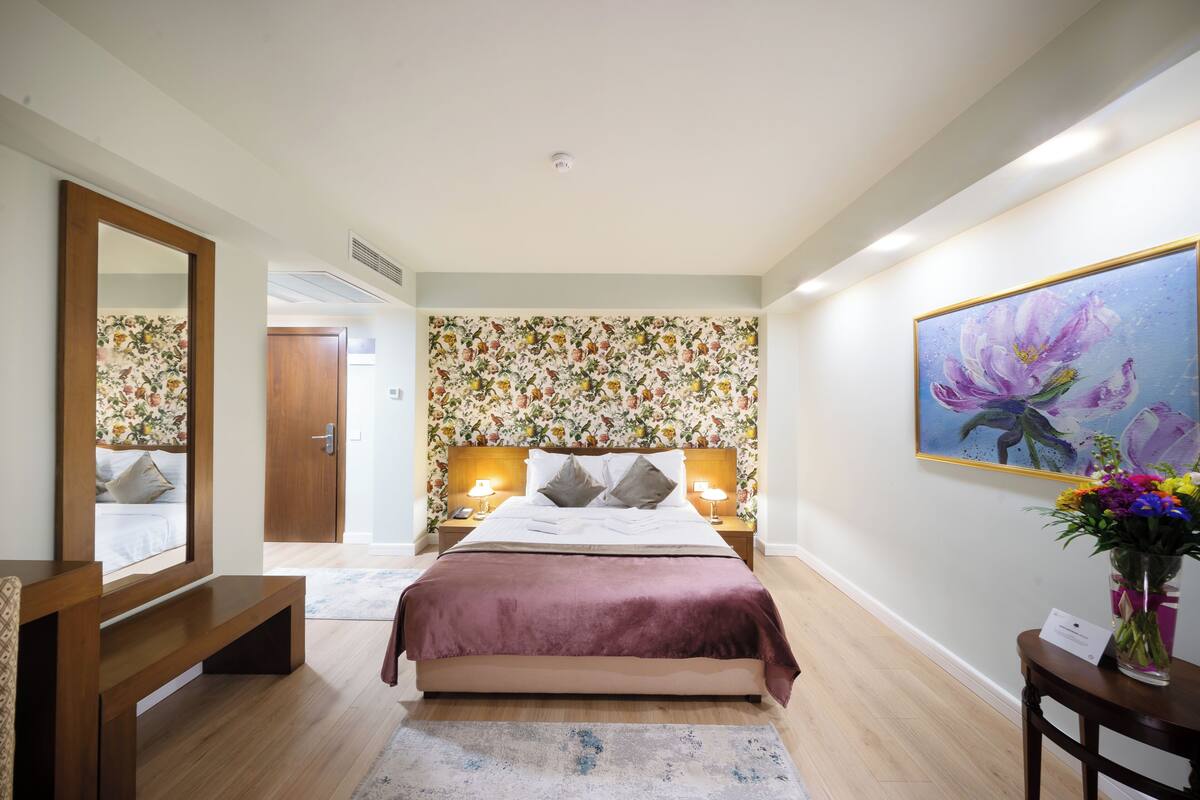 Une chambre à coucher avec un lit, des lampes de chevet, un miroir, un banc en bois, un papier peint à motifs floraux, un tableau et un vase avec des fleurs.