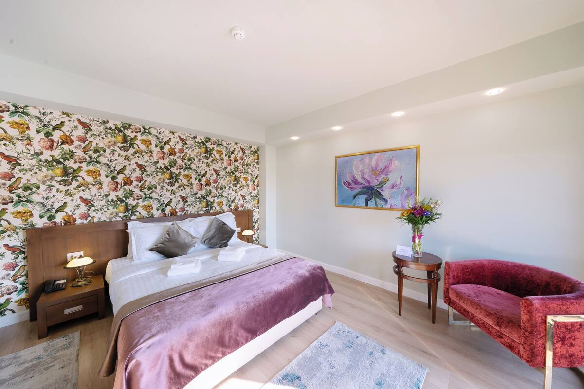 Une chambre à coucher avec un papier peint à motifs floraux, un lit recouvert d’une couverture violette, une chaise rouge et un tableau accroché au mur.