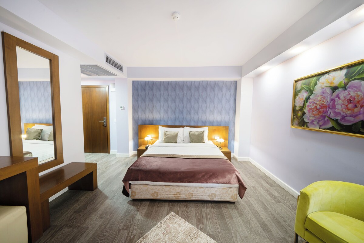 Una habitación de hotel con una cama, mesitas de noche, un espejo, un banco, una silla y un cuadro en la pared.