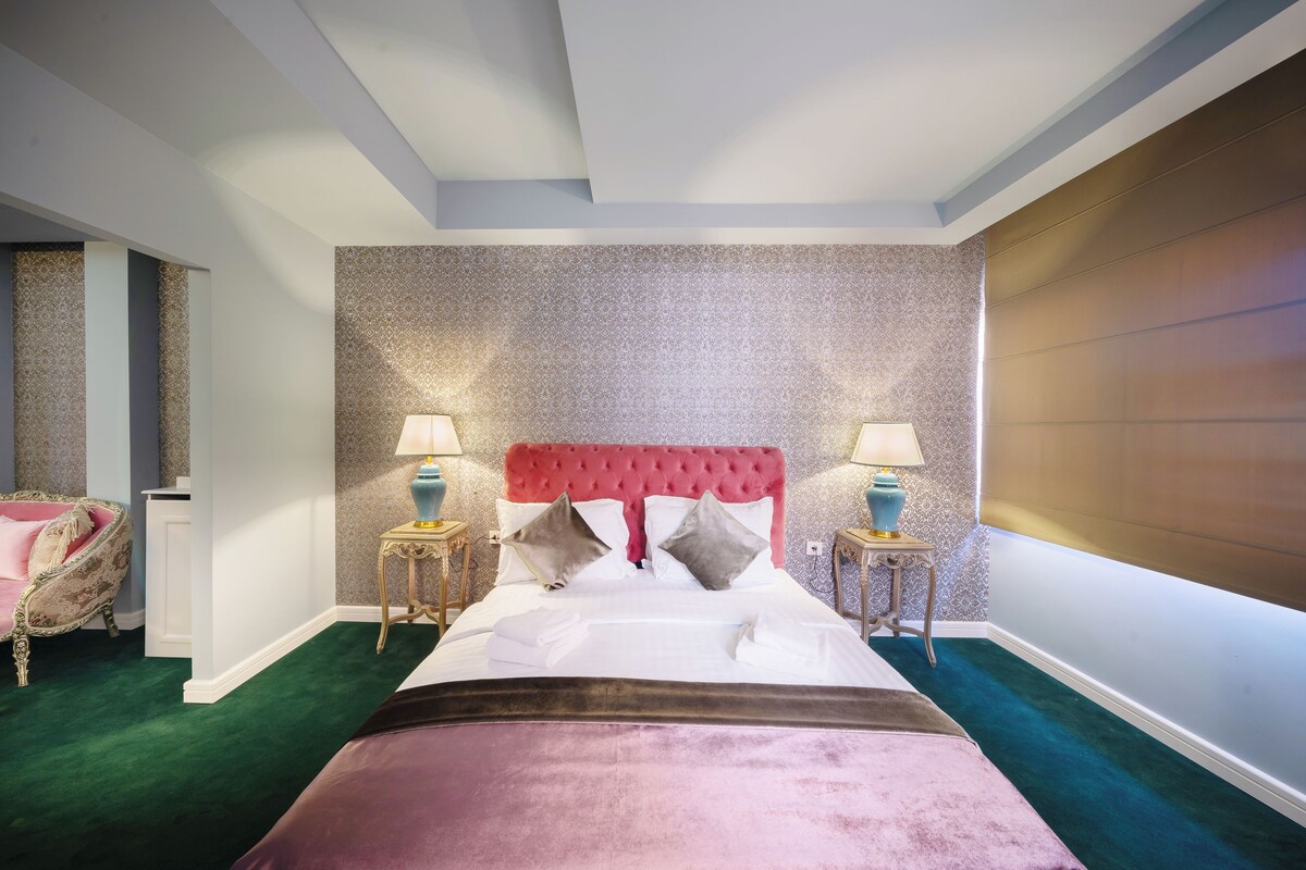 Habitación de hotel con una cama grande, dos mesitas de noche con lámparas, una silla decorativa y un papel tapiz estampado.