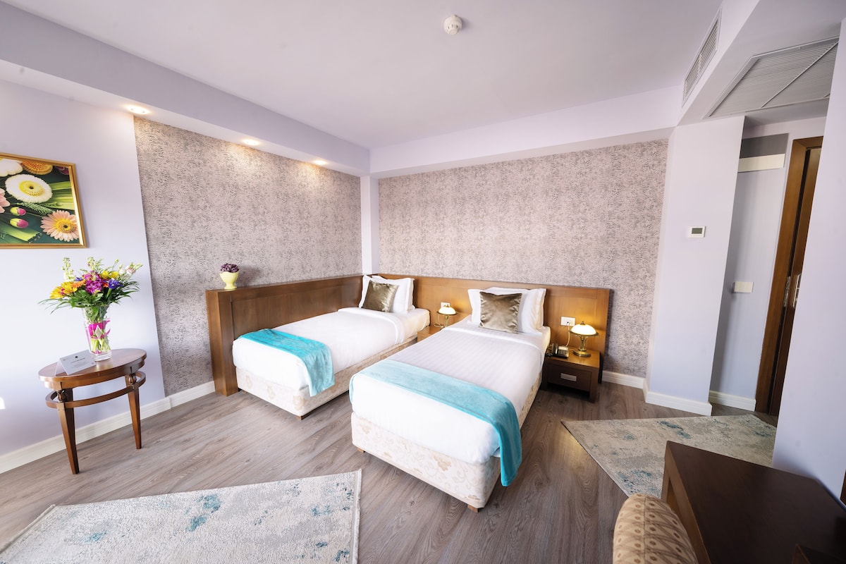 Une chambre d’hôtel avec deux lits, une petite table ornée d’un vase de fleurs et une table de chevet avec une lampe.