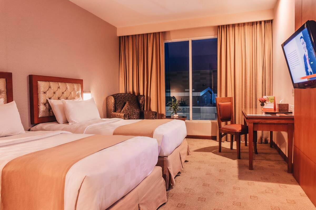 A hotel room with two beds, a desk, a chair, a television, and a large window with curtains.
