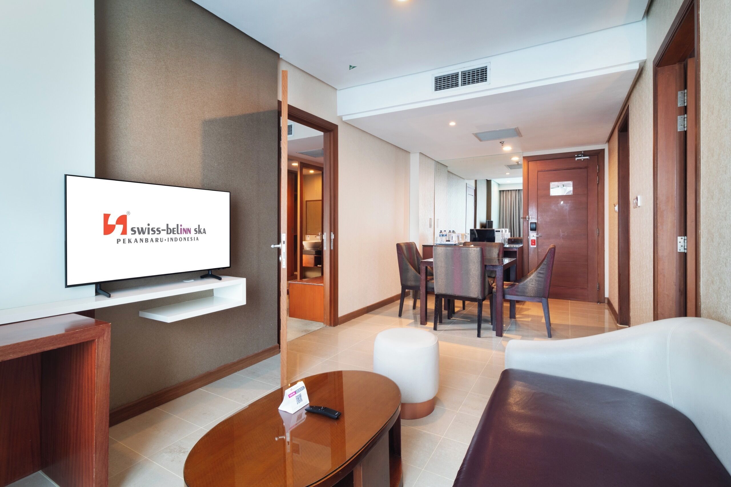 Habitación de hotel con televisor, una mesita de centro de madera, un sofá blanco, un área de comedor con una mesa y sillas, y una puerta de madera.