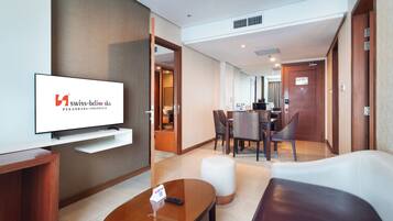 Habitación de hotel con televisor, una mesita de centro de madera, un sofá blanco, un área de comedor con una mesa y sillas, y una puerta de madera.