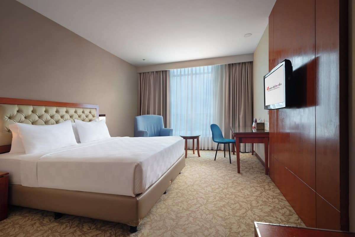 A hotel room with a large bed, a desk, two chairs, a television, and a window with curtains.