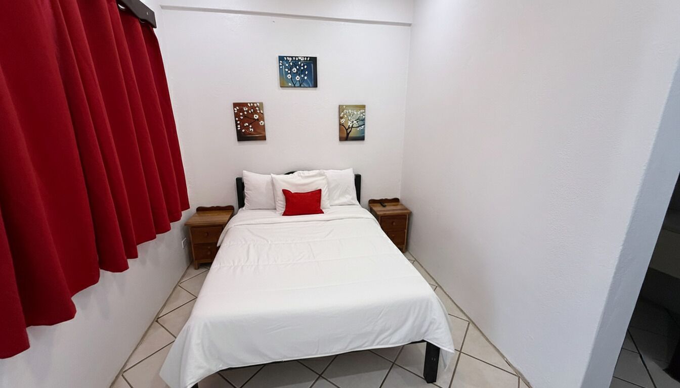 Quarto pequeno e bem organizado, com cama, duas mesas de cabeceira e cortinas vermelhas.