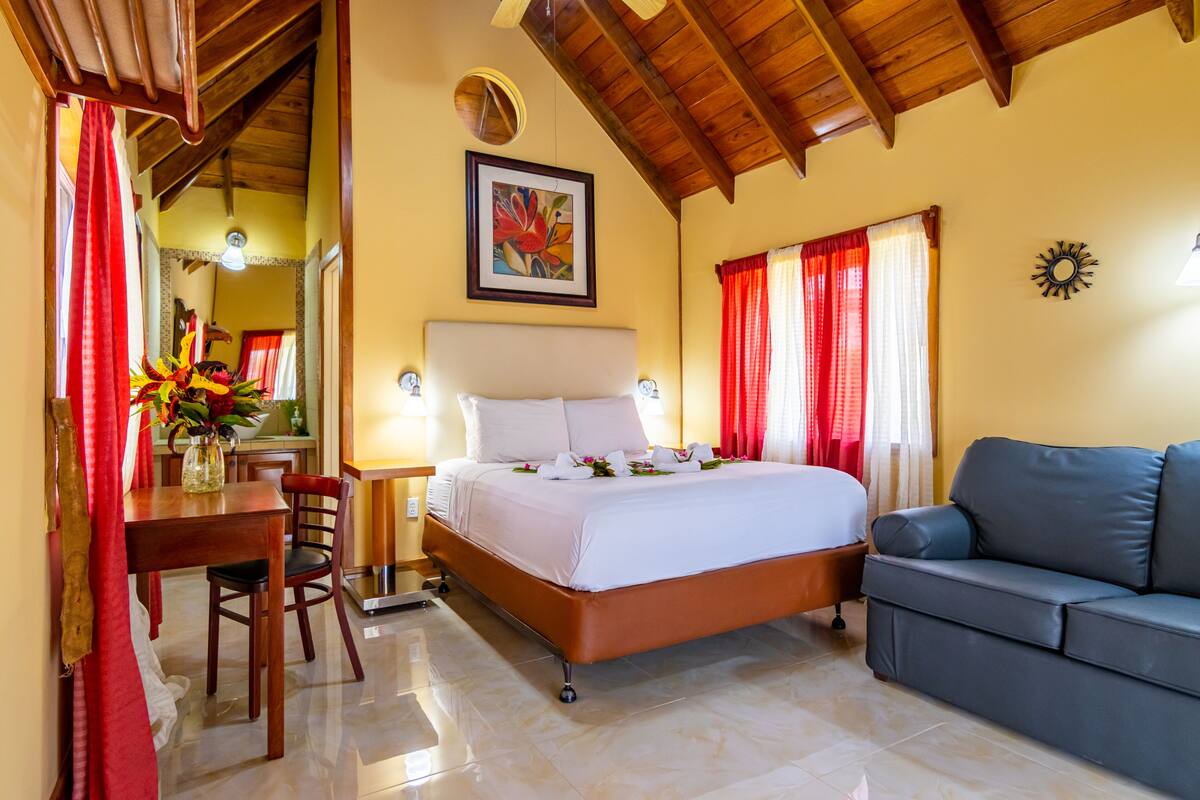 A hotel room with a bed, a sofa, a dining table with chairs, and a vase with flowers.