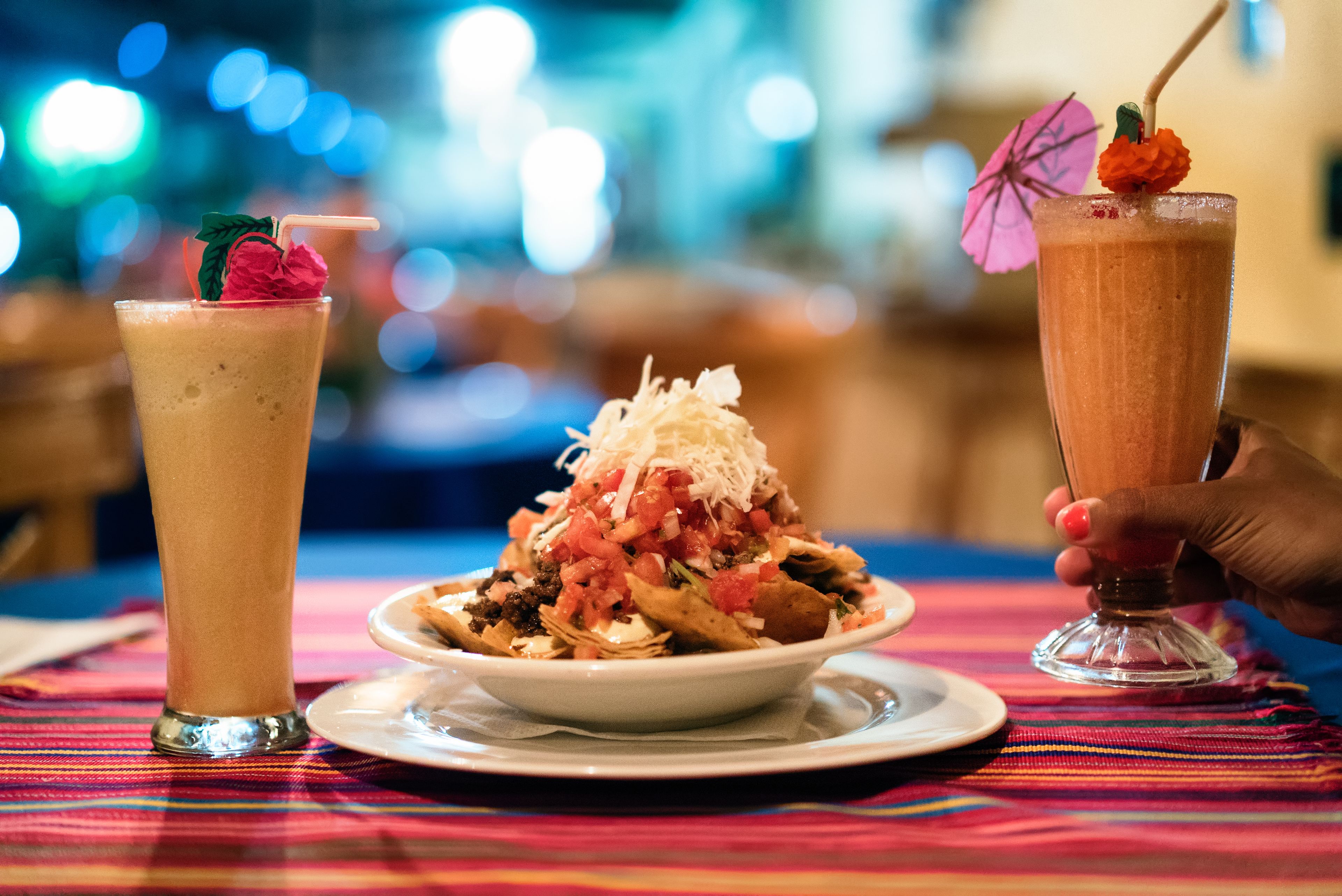 Una mesa con un mantel colorido, un tazón de nachos cubiertos de queso y tomates, y dos bebidas con pajitas y adornos.
