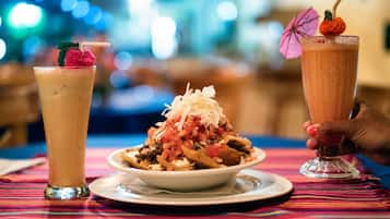 Uma mesa com toalha colorida, um prato de nachos coberto com queijo e tomates, e duas bebidas com canudos e enfeites.