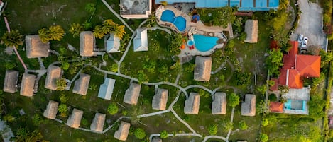 An aerial view of a resort with thatched-roof cottages, a swimming pool area, and a large building with a red roof.